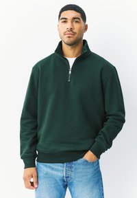 Tmavě zelená mikina s půlzipem, volným střihem, žebrovanými manžetami a pasem. Měkká textura, kombinovaná se světle modrými džínami.