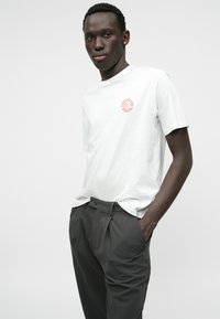 Camiseta blanca de manga corta con un logo circular en el lado izquierdo del pecho, combinada con pantalones gris plegados, de pie frente a un fondo blanco.