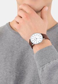 Silberne Uhr mit einem weißen Zifferblatt und schwarzen Markierungen, montiert an einem braunen Lederarmband, am Handgelenk getragen, mit einem sichtbaren grauen Pullover.