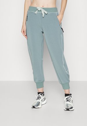 Person iført turkise joggerbukser med hvide syninger, elastiske manchetter, snøre i taljen og hvide sneakers med sorte detaljer.