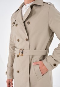 Cappotto beige con design a doppiopetto, dotato di bottoni marroni, cintura abbinata e tasche laterali. Tessuto con texture liscia.