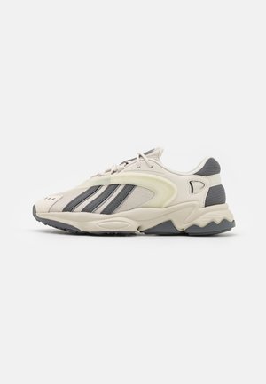 Sportieve beige en grijze sneaker met zwarte strepen, vetersluiting, gestructureerde zool en details van mesh-stof, gezien vanaf de zijkant.