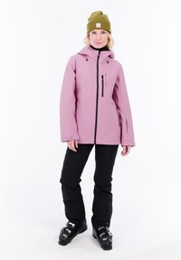 Woman wearing a green beanie, pink ski jacket, black ski pants, and black ski boots, standing on a white background.