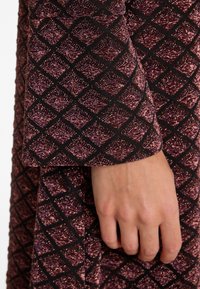 Long-sleeve garment with a textured, dark fabric featuring a diamond pattern in shades of purple and black, highlighting a shimmering effect.