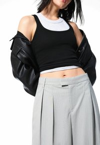 Schwarzes geripptes Tanktop, das über einem grauen Grafik-Tanktop getragen wird, kombiniert mit hellgrauen, gestreiften, plissierten Hosen und einer schwarzen Lederjacke.