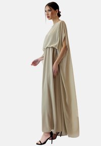 Beige, vloeiende maxi-jurk van lichtgewicht stof met brede mouwen en een elegante val. Gecombineerd met zwarte stilettohakken.