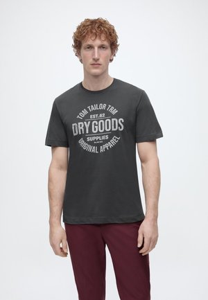 Νεαρός άνδρας με σγουρά κόκκινα μαλλιά, φορώντας μαύρο μπλουζάκι με το γράφημα «Dry Goods» και βυσσινί παντελόνι, στέκεται μπροστά σε απλό λευκό φόντο.