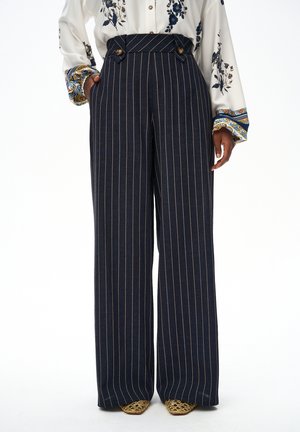Granatowe spodnie w paski z wysokim stanem i szerokimi nogawkami, zdobione guzikami, noszone z kwiecistą bluzką z długim rękawem i złotymi spiczastymi butami.