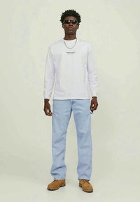 Man som bär svarta solglasögon, vit långärmad skjorta med logotyp, ljusblå jeans, beige stövlar och ett silverhalsband, som står mot en neutral bakgrund.