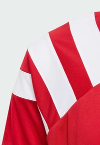 Rød og hvit sporty skjorte med diagonale hvite striper på rød stoff. Teksturen er glatt med en avslappet, atletisk passform.