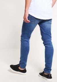 Jeans ajustados en denim azul medio con una textura suave, diseño de cinco bolsillos y detalles de costura sutiles. Combinados con zapatos negros sin cordones.