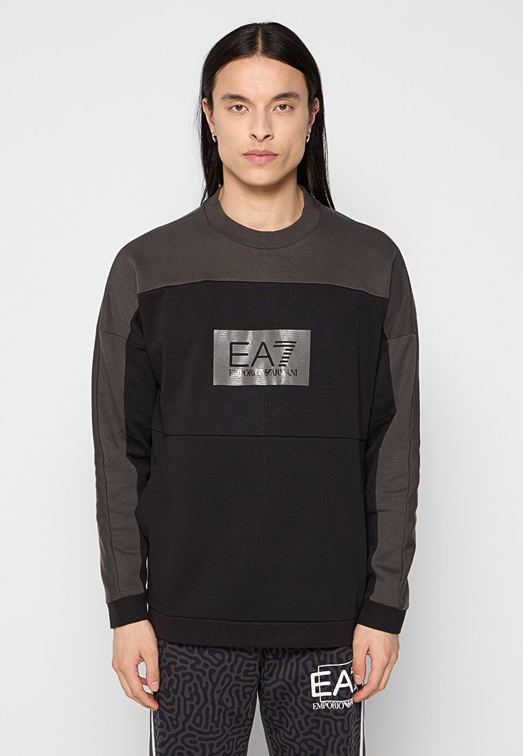Felpa a maniche lunghe nera e grigia scura con una texture a coste, caratterizzata da un logo EA7 metallico sul davanti. Design semplice e moderno.