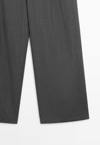 Pantalon large gris foncé à fines rayures montrant la partie inférieure de la jambe sur fond blanc.