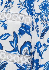 Blaues Blumenmuster auf weißem Stoff mit Spitzenverzierungen. Enthält große Blumen, Blätter und kreisförmige Spitzenakzente entlang der Nähte.