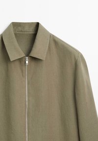 Giacca leggera verde oliva con chiusura a zip, colletto piatto e maniche lunghe. Presenta una texture sottile e un design pulito e minimalista.