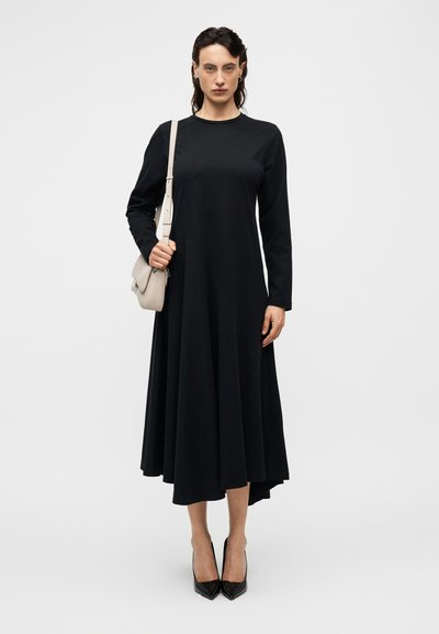 Μαύρο φόρεμα με μακριά μανίκια, φούστα με φούντα και ασύμμετρη λοξή βάση, κατασκευασμένο από λείο ύφασμα. Συνδυάζεται με ανοιχτό γκρι τσάντα ώμου και μαύρα ψηλοτάκουνα.