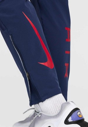 Pantalones deportivos de color marino con el logotipo y la tipografía de Nike en rojo, bolsillos laterales con cremallera y puños ajustados. Combinados con zapatillas grises y moradas.