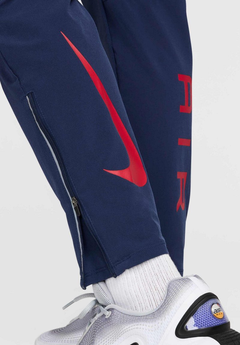 Granatowe spodnie sportowe mają czerwone logo i napisy Nike, boczne kieszenie na zamek oraz zwężane mankiety. W komplecie z szaro-fioletowymi sneakersami.