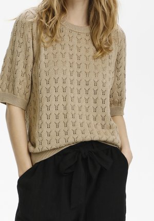Femme portant un pull en tricot beige à manches courtes avec de petits motifs ajourés et un pantalon noir taille haute noué avec un nœud en tissu.