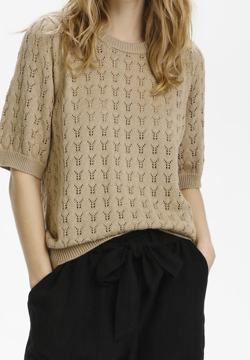 Femme portant un pull en tricot beige à manches courtes avec de petits motifs ajourés et un pantalon noir taille haute noué avec un nœud en tissu.
