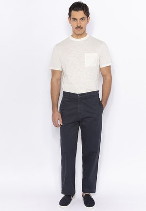 Light gray short-sleeve t-shirt with a front pocket, paired with dark navy pants. Model wearing slip-on navy shoes. Simple, casual outfit.