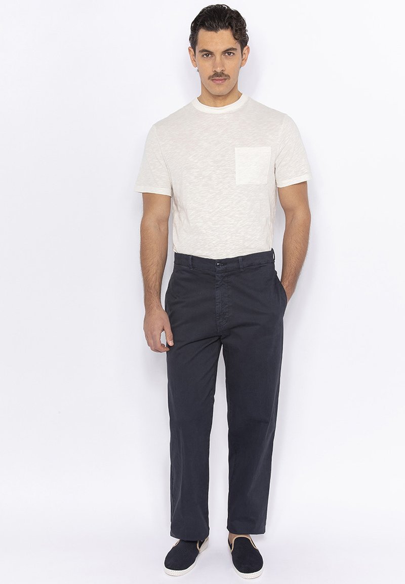 Lichtgrijs t-shirt met korte mouwen en een voorzak, gecombineerd met donkerblauwe broek. Model draagt navy instapschoenen. Eenvoudige, casual outfit.