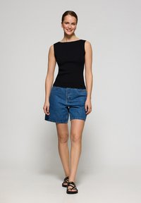 Svart ärmlös topp i kombination med högmidjade blå denimshorts. Modellen har på sig svarta sandaletter; enkel, ren design utan synliga mönster.