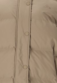 Beige puff jakke med en glat tekstur, runde knapper og et subtilt logo. Designet inkluderer vandret syninger for isolering.