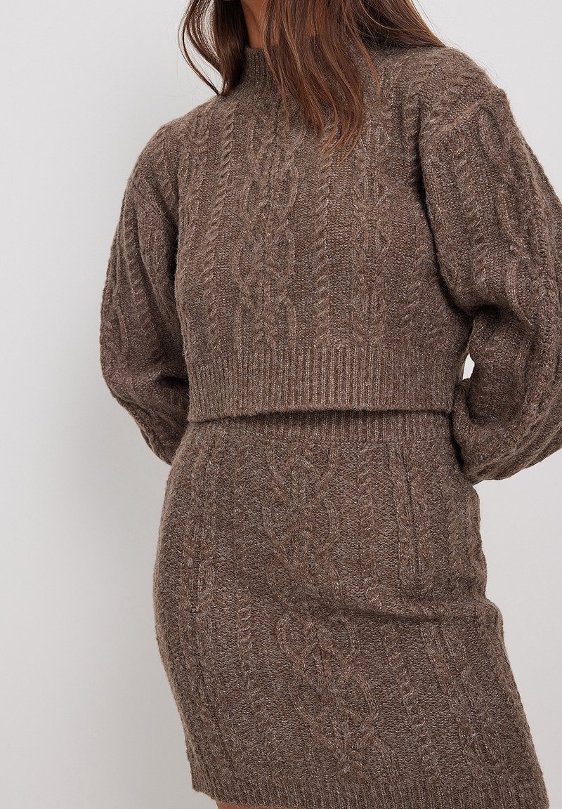 Femme portant un pull en maille torsadée marron assorti et une jupe avec des ourlets côtelés, visible de la moitié du torse jusqu’au milieu des cuisses sur fond uni.