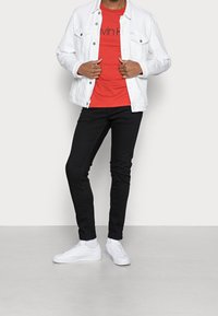 Giacca di jeans bianca sopra una t-shirt rossa di Calvin Klein, abbinata a jeans skinny neri e sneaker bianche. Design semplice, forma aderente.