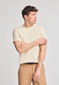 Licht beige T-shirt met korte mouwen en een ronde halslijn, voorzien van een subtiele logo-detail. Soepele stof met een relaxte pasvorm, gecombineerd met tan kleurige broek.