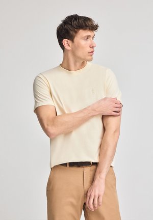 T-shirt corto beige chiaro a maniche corte con scollatura rotonda e un sottile dettaglio del logo. Tessuto morbido con una vestibilità rilassata, abbinato a pantaloni beige.