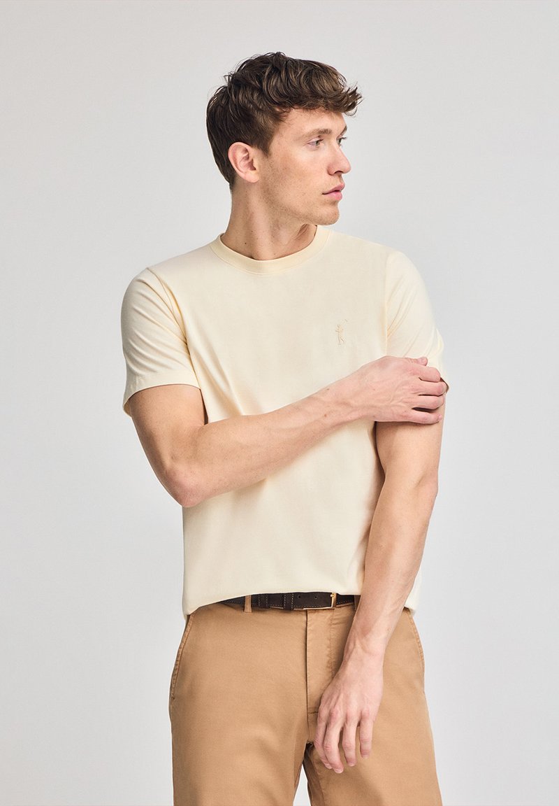 Licht beige T-shirt met korte mouwen en een ronde halslijn, voorzien van een subtiele logo-detail. Soepele stof met een relaxte pasvorm, gecombineerd met tan kleurige broek.