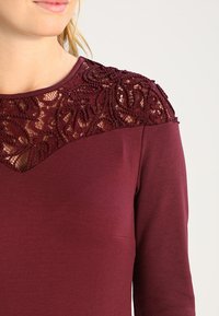 Mujer vistiendo una blusa de manga larga color burdeos con intrincados detalles de encaje en la parte superior del pecho y los hombros.