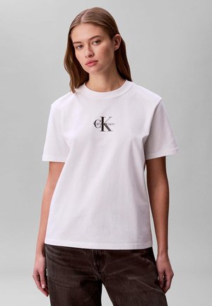 Giovane donna con indosso una t-shirt bianca Calvin Klein con logo, abbinata a pantaloni scuri, in piedi su uno sfondo grigio neutro.