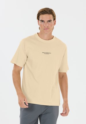 Jeune homme aux cheveux courts châtain clair, portant un t-shirt beige avec le texte "Quality Essentials" et un pantalon gris, debout sur un fond blanc uni.