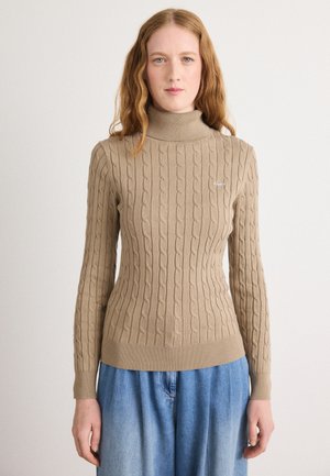 Beige kabelstrikket turtleneck genser med ribbestrikkede mansjetter og hem, kombinert med løse lyseblå denimbukser. Enkel brodert logo detaljer.