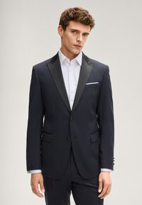 Giacca da smoking blu marina con colletto a scialle nero in raso, chiusura con un solo bottone, due tasche frontali e un fazzoletto bianco nel taschino.