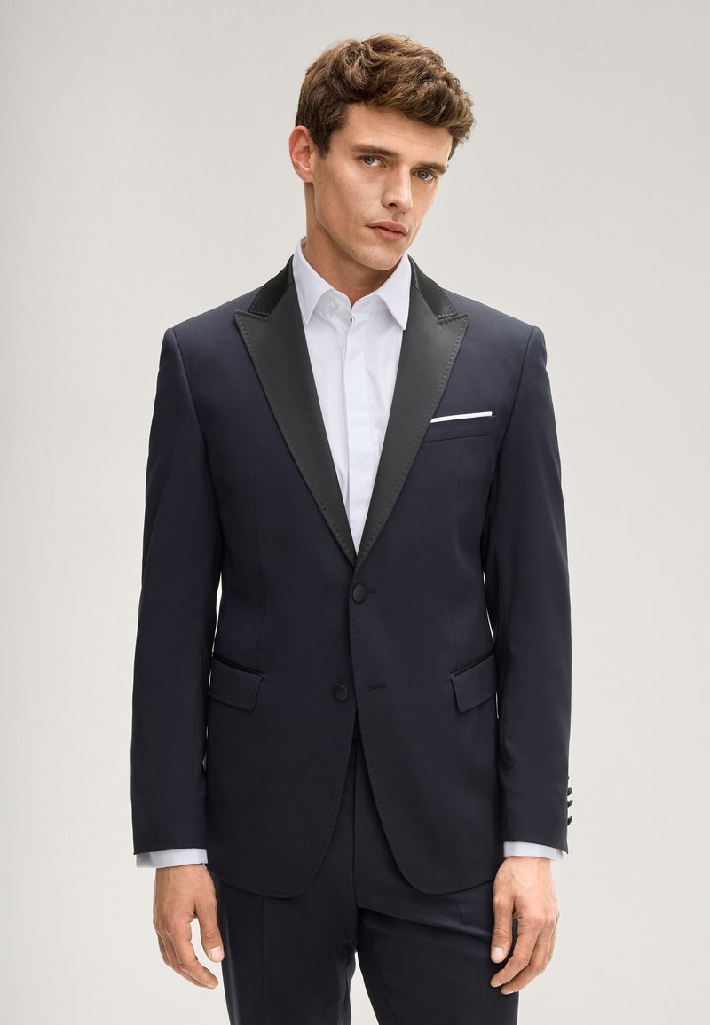 Giacca da smoking blu marina con colletto a scialle nero in raso, chiusura con un solo bottone, due tasche frontali e un fazzoletto bianco nel taschino.