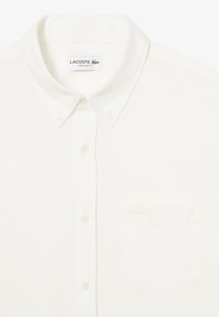 Weißes Baumwollhemd mit klassischem Kragen, ausgestattet mit einer linken Brusttasche und dem Lacoste-Logo in tonal gestickter Ausführung.