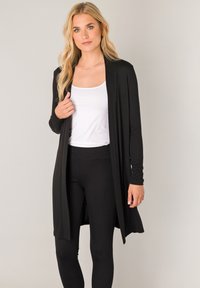 Zwarte lange cardigan gemaakt van zachte stof, met lange mouwen en een open voorkant. Gecombineerd met een wit topje en zwarte legging.