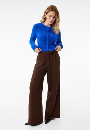 Vrouw die een felblauwe, pluizige, geknoopte cardigan rechtzet, draagt hoge taille wijde bruine broeken en zwarte puntige schoenen, staand op een witte achtergrond.