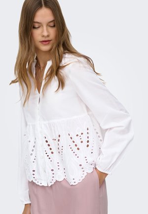 Kvinde med langt brunt hår iført en hvid knappet bluse med hulbroderi og lyserøde bukser, kigger ned med den ene hånd i lommen.