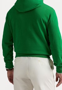 Grön hoodie i bomullsblandning med avslappnad passform, utrustad med huva, ribbade muddar och fåll, kombinerad med cremefärgade byxor som visar knappdetaljer.