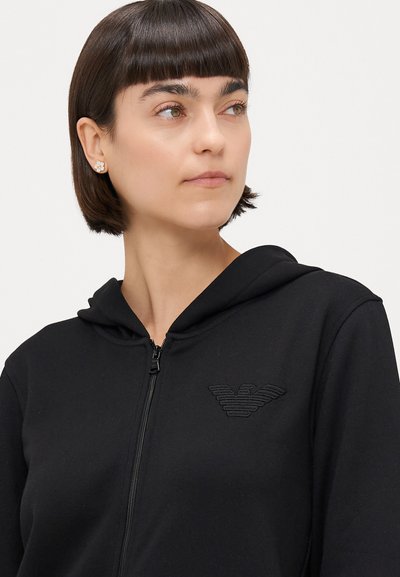 Črn zip-up pulover s kapuco; ima embosirano logo na prsih. Mehka tkanina z gladko teksturo.