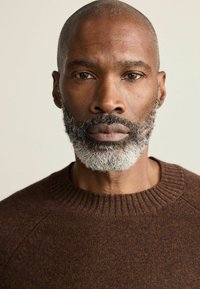 Brauner Pullover mit Rippenausschnitt, getragen von einem Mann mit einer glatzen Kopf und grauem Bart. Der Stoff wirkt strukturiert und warm.