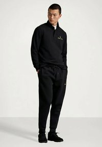 Άνδρας που στέκεται με τα χέρια στις τσέπες, φορώντας μαύρο μακρυμάνικο πόλο, συνδυασμένα μαύρα joggers και μαύρα αθλητικά παπούτσια, σε απλό φόντο.