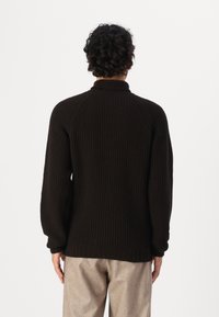 Donkerbruine geribde trui met een hoge kraag en raglanmouwen, met een relaxte fit en een textuurpatroon, gedragen over beige broek.
