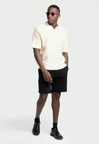 Homem com polo de malha de manga curta beige, calções pretos, meias pretas, sapatos pretos brilhantes e óculos de sol, a caminhar com uma mão no bolso.