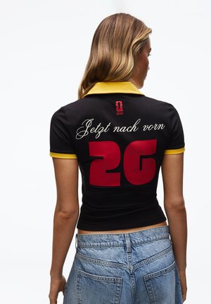 Femme vue de dos portant une chemise noire avec un col et des poignets jaunes, texte rouge "2G" et écriture blanche "Jetzt nach vorn" dans le dos.